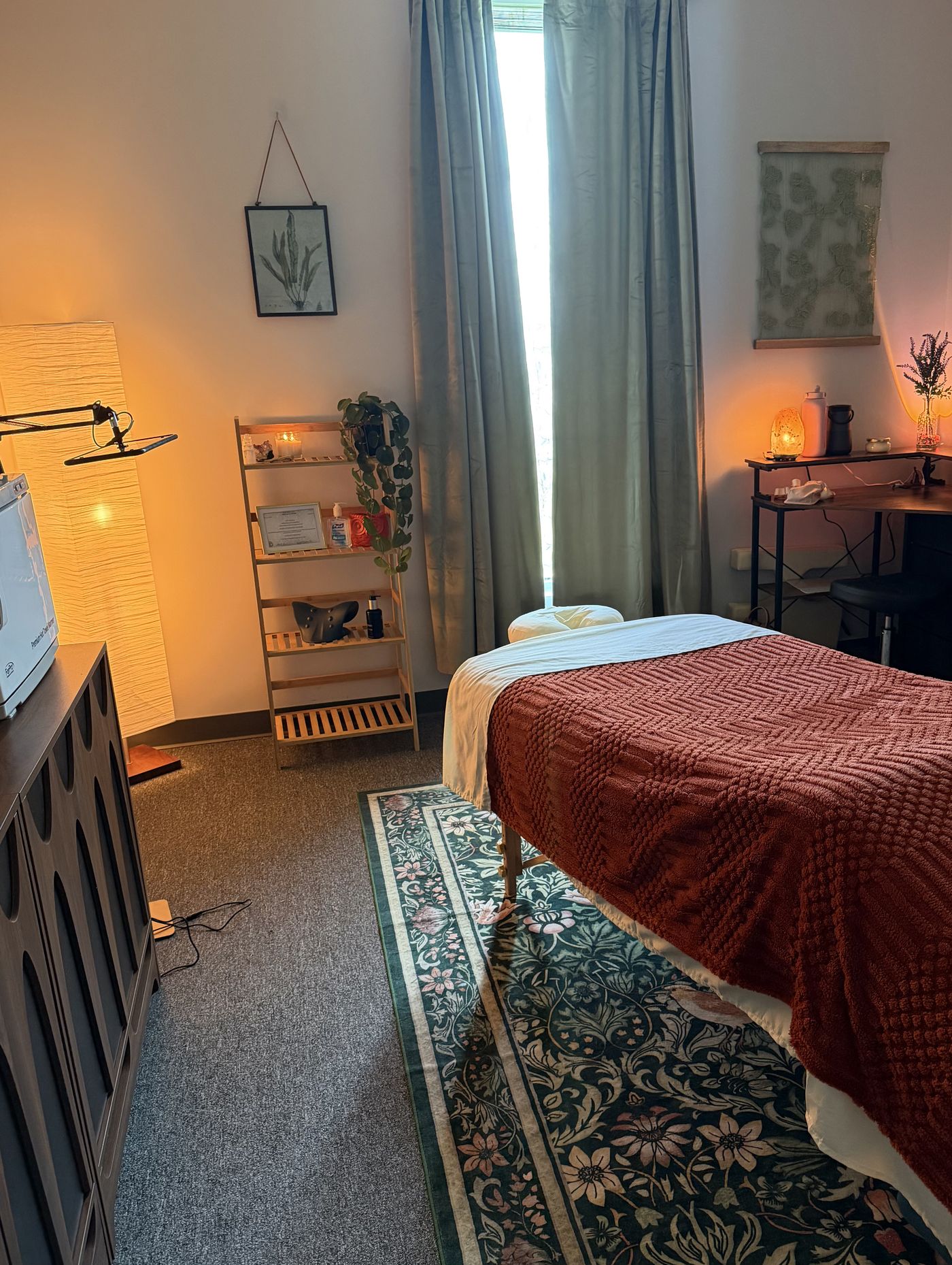 Inside the Luma studio: a softly-lit massage room with sage curtains, a terracotta knit blanket, plants, and warm lamp light.